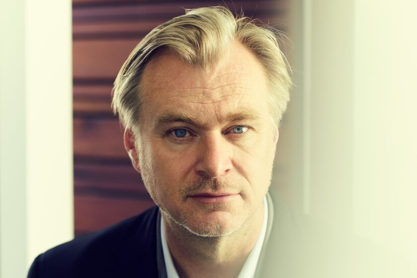 A blond man in a dark blazer looks through a reflective panel of glass